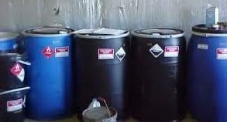 Image shows a storage area with multiple blue and black 55-gallon drums labeled hazardous material.