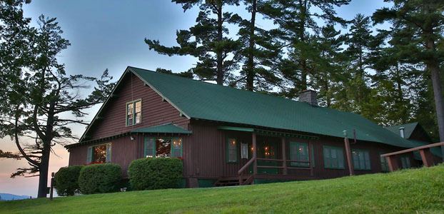 Brown wooden lodge with a green metal roof with a mountain setting in the background.
