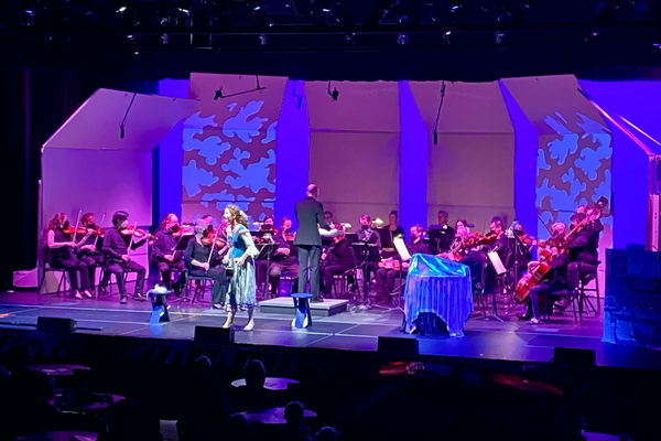 An orchestra, conductor, and singer perform on a stage lit with blue and purple lights