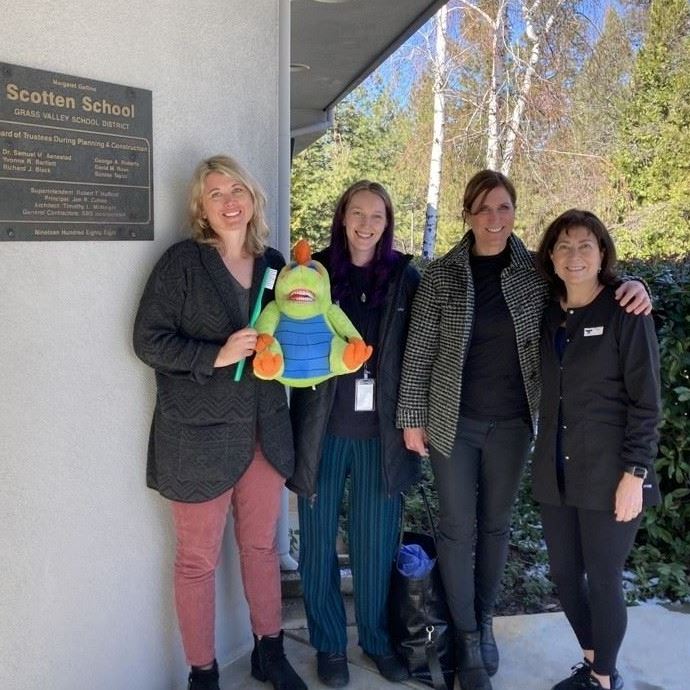 Four people stand near a Scotten School sign. One person holds a green stuffed animal and a large gr