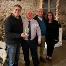 Four people stand together smiling, one holds a colorful blown glass award.