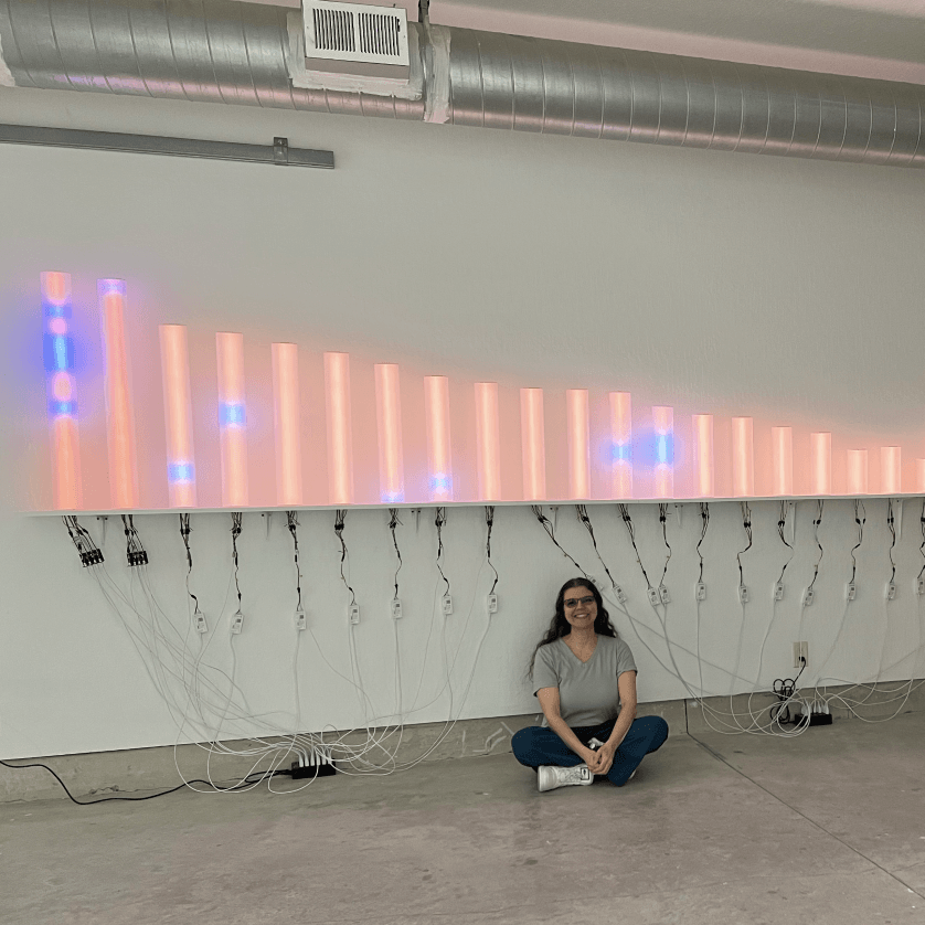 Artist Rachel Rein sits on the floor underneath an orange LED light sculpture