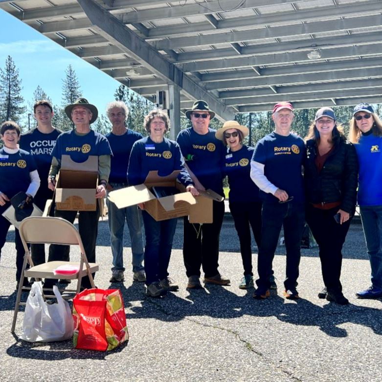 A group of people in blue Rotary t-shirts stand in the parking lot of the Rood Center holding boxes