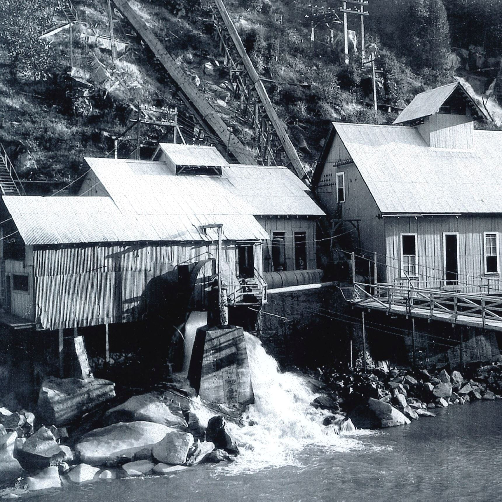 Two tin buildings sit along the water's edge below a steep vegetated slope. 