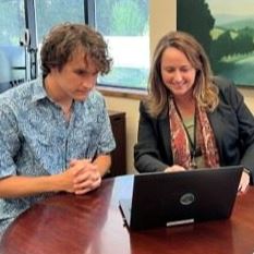 Logan van den Berg and Alison Lehman work together on a laptop at a table in teh CEO's office.