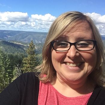white woman with blonde hair and glasses smiles with the mountains and blue sky in the background