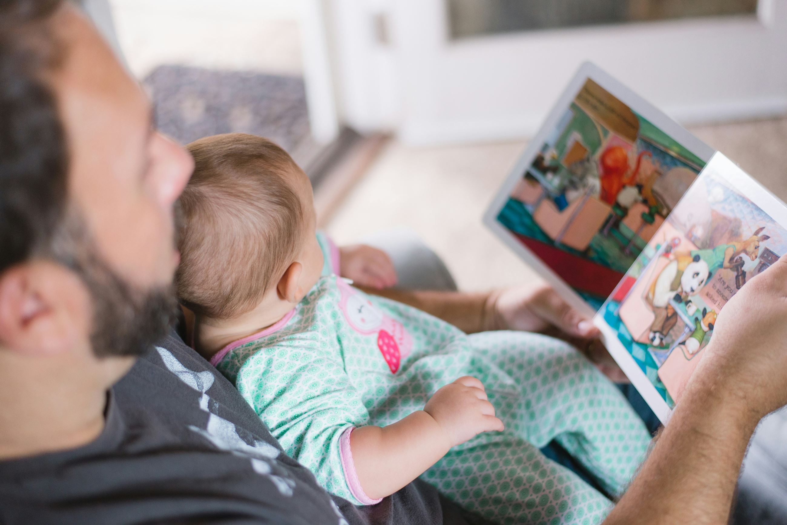 Photograph over the shoulder of a white man reading to a white baby seated in his lap.