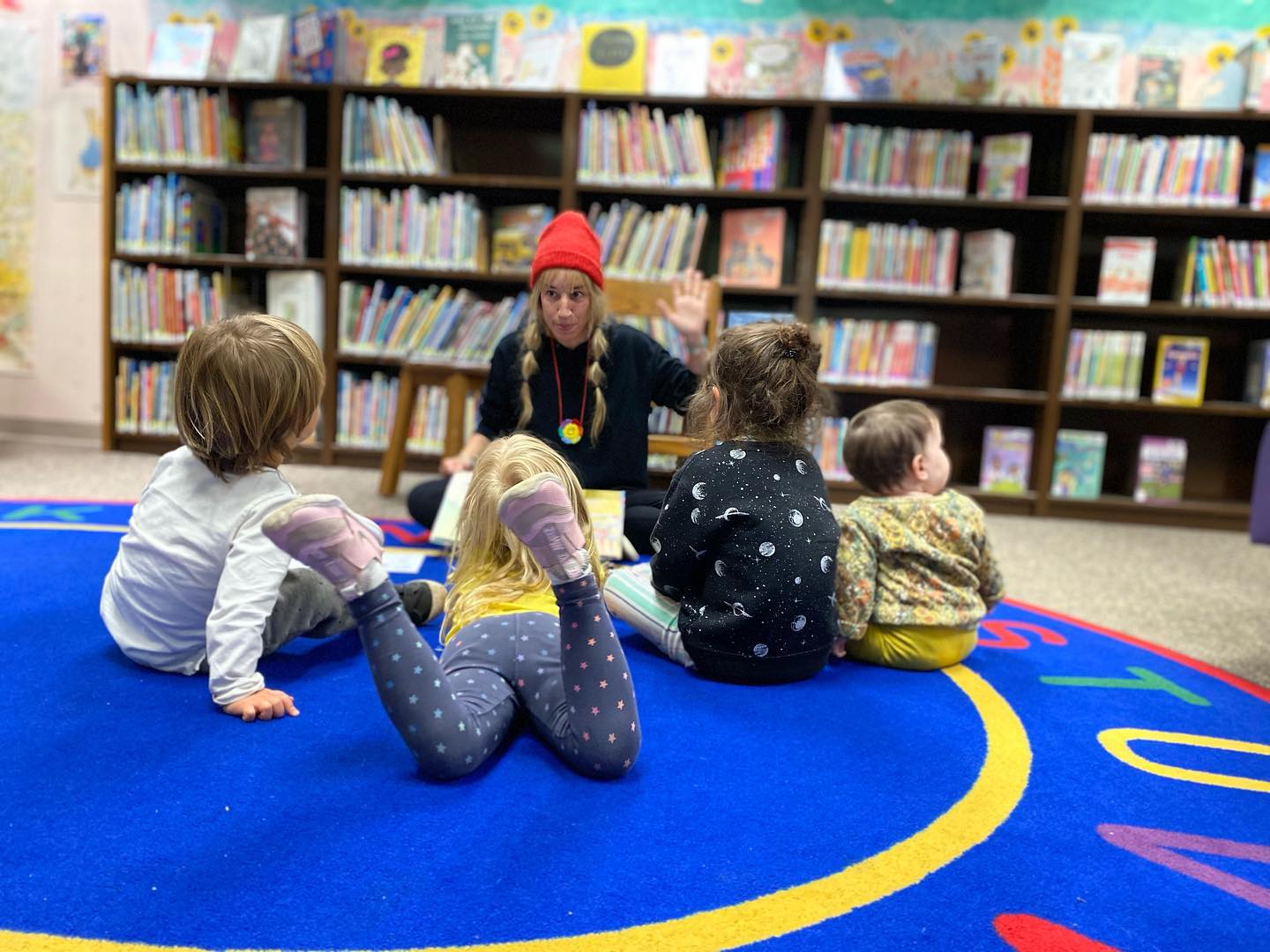 Children sitting on a carpet facing a storyteller with books in the background
